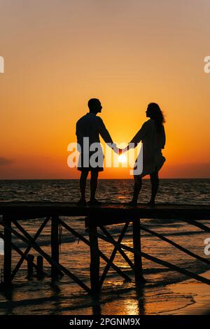 Silhouette de couple tenant les mains et debout sur la jetée au lever du soleil Banque D'Images