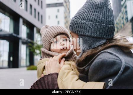 Femme portant un chapeau en tricot embrassant son fils devant le bâtiment Banque D'Images