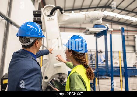 Les ingénieurs portant un casque de sécurité discutant devant le bras robotisé à l'usine Banque D'Images