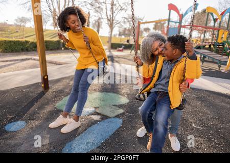 Bonne femme regardant la mère poussant petit-fils sur balançoire sur le terrain de jeu Banque D'Images