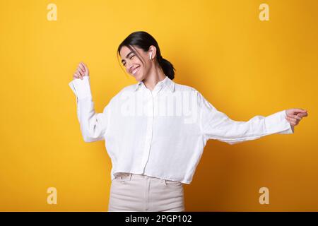 Une femme heureuse qui porte des écouteurs intra-auriculaires sans fil pour écouter de la musique et danser sur fond jaune Banque D'Images