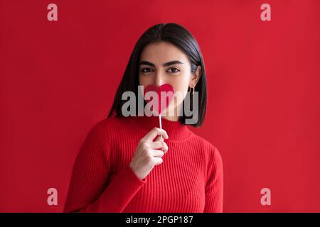 Jeune femme tenant un lollipop en forme de coeur sur la bouche contre fond rouge Banque D'Images