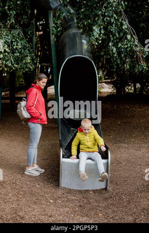 Curieux petit garçon dans des vêtements chauds jouant sur le toboggan de l'aire de jeux avec sa maman. La mère caucasienne se tient et prendre soin de petit garçon jouer diapositive dans le plaisir Banque D'Images