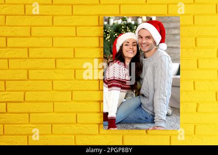 Toile avec photo imprimée Happy couple portant des chapeaux de père Noël sur le mur de brique jaune, espace pour le texte Banque D'Images