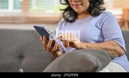 Image rognée d'une heureuse femme asiatique de 60 ans à la retraite utilisant son téléphone sur un canapé dans le salon. Banque D'Images