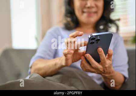 Image rognée d'une heureuse femme asiatique de 60 ans à la retraite utilisant son téléphone sur un canapé dans le salon. discussion, défilement sur le téléphone, lecture d'un sur Banque D'Images