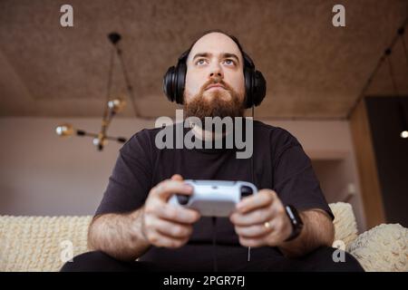 Style de vie d'un jeune joueur masculin jouant sur la console avec des casques à tenir le jeu ontroller dans ses mains. Banque D'Images