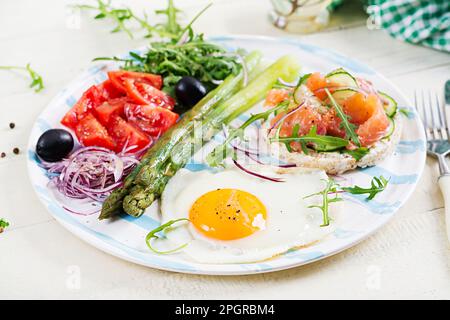 Petit déjeuner cétogène. Petit déjeuner. Œuf frit, pains croustillants au saumon, asperges vertes, tomates fraîches et oignons rouges sur plaque blanche. Banque D'Images