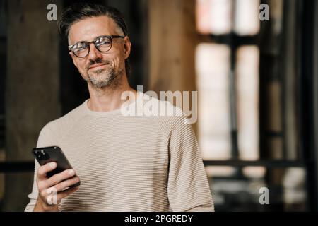 Homme blanc d'âge moyen dans des lunettes souriant à l'appareil photo et tenant le téléphone portable à l'intérieur Banque D'Images