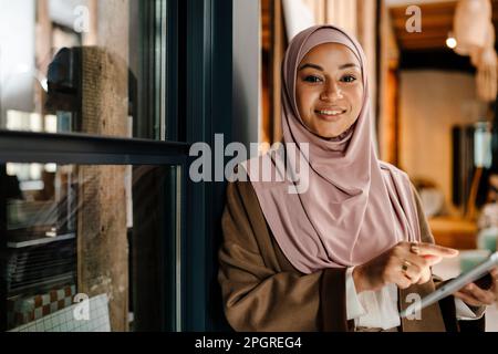 Femme arabe gaie portant le hijab travaillant sur une tablette tout en étant debout dans un bureau moderne Banque D'Images