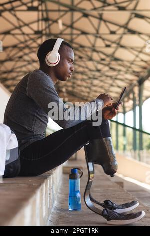 Un athlète masculin en forme de compétition mixte dans un stade sportif en plein air, assis dans les tribunes, avec un casque et des lames de course à pied sur un smartphone. Banque D'Images