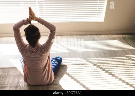 Vue arrière de la jeune femme caucasienne avec les bras levés méditant tout en étant assise sur le sol dans la salle de séjour. Espace de copie, non modifié, yoga, zen, style de vie, gagnez Banque D'Images