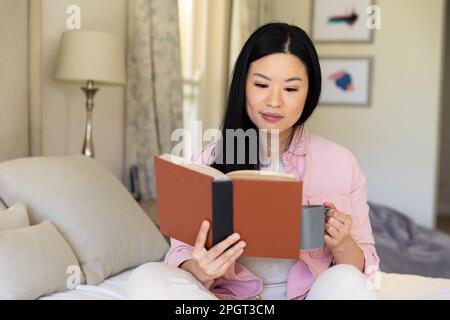Femme asiatique souriante assise sur son lit lisant un livre et buvant du café pendant la journée. Style de vie, temps libre et détente. Banque D'Images