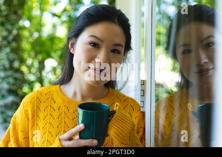 Portrait d'une femme asiatique heureuse portant un chandail jaune debout près de la fenêtre souriant et buvant du café. Vie domestique, temps libre et détente. Banque D'Images