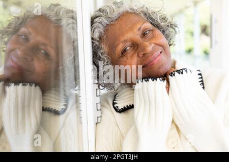 Gros plan d'une femme afro-américaine souriante avec les mains sur le menton penchées sur la fenêtre du balcon. Non modifié, style de vie, portrait, réflexion et retraité Banque D'Images