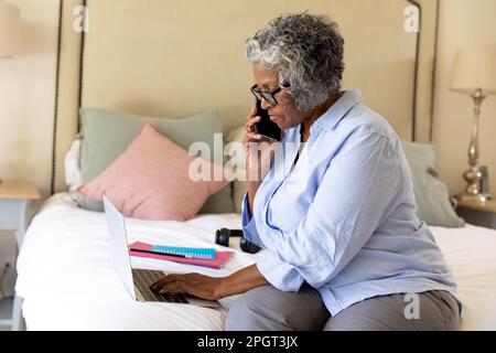 Femme d'affaires afro-américaine spécialisée, qui parle sur un téléphone portable et utilise un ordinateur portable au lit. Espace de copie, non modifié, professionnel, travail à domicile, Banque D'Images