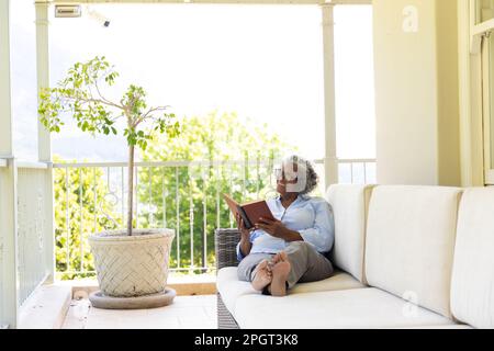 Pleine longueur de bonne femme afro-américaine senior lisant le livre tout en se relaxant sur le canapé dans le balcon. Espace de copie, non modifié, style de vie, passe-temps, lunettes Banque D'Images