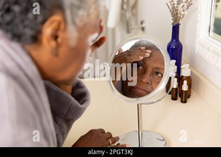 Une femme afro-américaine âgée touche son visage et vérifie les imperfections, les rides dans le miroir à la maison. Sans altération, soins du corps, soins de la peau, vieillissement, réflexion Banque D'Images