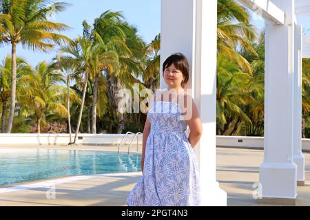 Portrait d'une femme heureuse vêtue d'été debout près de la colonne blanche sur la piscine et le fond des palmiers à noix de coco. Vacances dans un complexe tropical Banque D'Images
