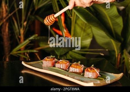 Gros plan d'une main humaine qui verse du miel sur une assiette de sushis fraîchement préparés Banque D'Images