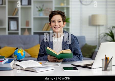 Portrait d'un jeune garçon d'école à la maison, un garçon avec des bretelles sur les dents atteint et regarde l'appareil photo, l'adolescent fait des devoirs à la maison et étudie à distance, en utilisant un ordinateur portable parmi les livres. Banque D'Images
