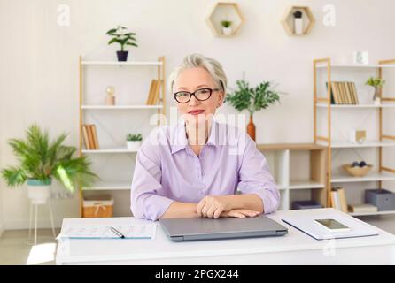 Heureuse belle femme d'affaires mûre et prospère assise à son bureau dans le bureau Banque D'Images