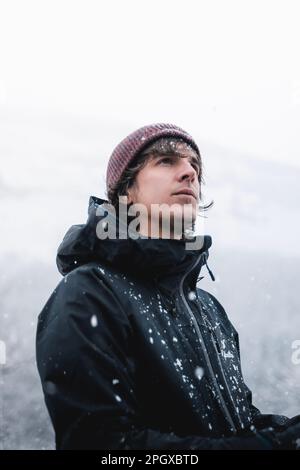 Un jeune homme avec un bonnet rouge en laine et une veste noire sous les flocons de neige en hiver. Banque D'Images