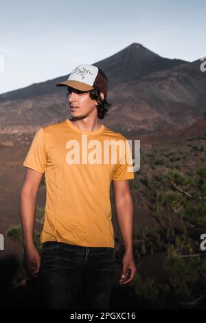 Jeune homme avec de longs cheveux et une casquette de camion de soleil avec chemise jaune dans un paysage de montagne au coucher du soleil à ténérife. Banque D'Images