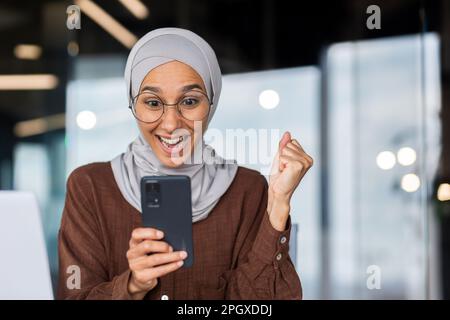 Gros plan sur une femme heureuse et prospère sur le lieu de travail dans un bureau moderne, une femme d'affaires dans le hijab et des lunettes lisant les bonnes nouvelles au téléphone, tenant joyeusement le geste de main vers le haut de triomphe et de réalisation. Banque D'Images