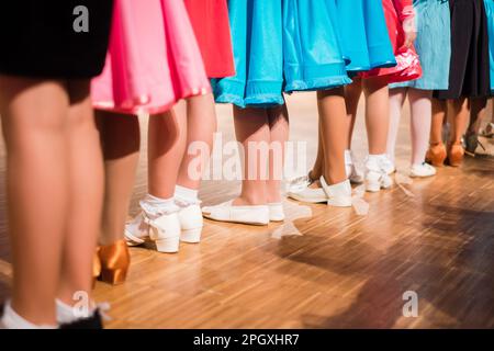 Jeunes filles danseuses de salle de bal debout dans une rangée avant la compétition pour débutants. Robes colorées et différentes chaussures sur parquet. Banque D'Images