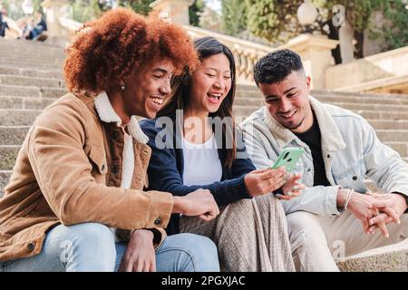 Groupe de trois jeunes amis multiraciaux souriant et s'amusant à l'aide d'une application de réseaux sociaux sur smartphone. Des adolescents heureux regardant des vidéos sur le smartphone assis dans les escaliers à l'extérieur. communication. Photo de haute qualité Banque D'Images