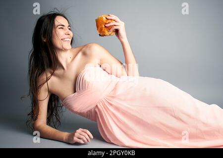 Femme en robe de bal rose de bal de bal de proéminent sur fond de papier gris sans couture tenant un hamburger Banque D'Images