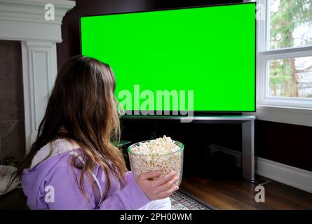 Jeune femme montres projecteur, TV, films avec pop-corn en soirée grande télévision verte écran fille est assis sur le sol près de la cheminée sur la moquette dans la veste violette portant des pantalons blancs est regarder la publicité de film Banque D'Images