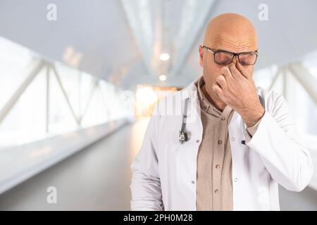 portrait d'un homme caucasien âgé médecin à l'hôpital. Arrière-plan flou. Frottant le nez et les yeux avec sa main. Homme stressé. Banque D'Images