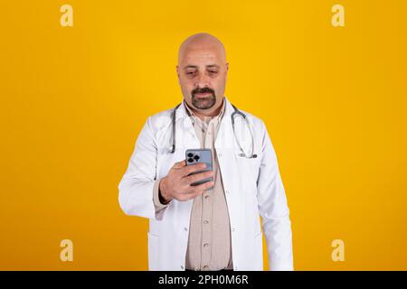 Portrait d'un médecin caucasien utilisant un smartphone. Arrière-plan jaune isolé, espace de copie. Médecin professionnel de l'hôpital chauve d'âge moyen. Banque D'Images