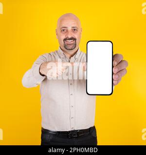 homme d'affaires mûr debout sur fond jaune et pointant le smartphone. Regarder l'appareil photo, sourire. Site Web, maquette de placement d'application. Banque D'Images