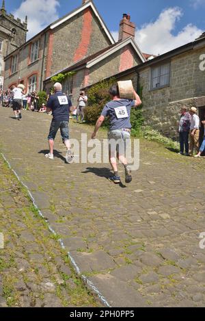 Les concurrents de la course du fromage masculin portent leurs fromages sur Gold Hill, Shaftesbury, lors du festival de la gastronomie et des boissons de la ville, le 8th mai 2022. Banque D'Images