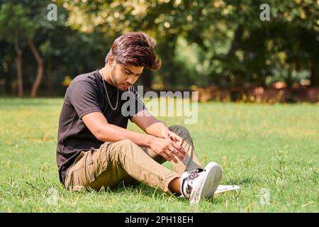 Beau portrait de jeune indien en t-shirt noir et chaîne de cou argent assis sur une pelouse verte dans un parc public, portrait d'homme hindou. Beau indien Banque D'Images