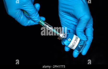 Main dans des gants médicaux bleus tenant un flacon en verre avec une étiquette de vaccin, fond noir Banque D'Images