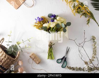 Faire bouquet fleuriste lieu de travail. Arrangement de fleurs dans un magasin de fleurs. Bouquet de printemps de fleurs jaunes. Iris, tulipes, mimosa et eucalyptus. Bouton tulipe. Fond floral. Iris violet. Table blanche Banque D'Images