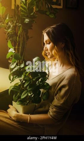 Une belle douce jeune femme blonde est assise à la maison avec une plante en pot dans ses mains lors d'une journée ensoleillée. Jardinage à la maison et confort. Plantes d'intérieur. Ce Banque D'Images