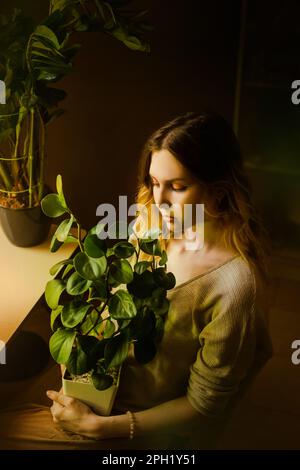Portrait d'une belle jeune blonde en vêtements beiges, qui est assise avec une fleur verte en pot dans ses mains à la maison. Amour des plantes. Jardinage à la maison Banque D'Images