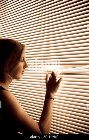 une femme blonde se tenant près d'une fenêtre avec des stores et regardant à l'extérieur Banque D'Images