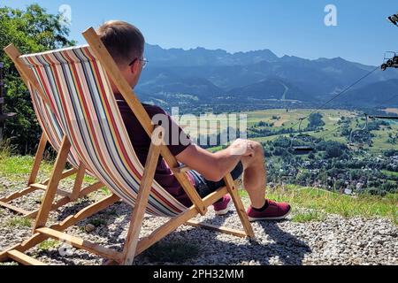 un jeune homme est assis dans un transat avec son dos à la caméra. donne en arrière, se détend, bénéficie de la vue sur les montagnes par une journée ensoleillée Banque D'Images