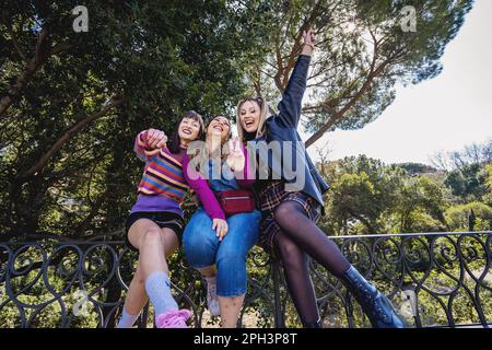 Trois femmes caucasiennes en 90s tenues décontractées vintage, assises sur une balustrade de parc, applaudissent et chantent ensemble. Ils gestent avec leurs mains, avec un t Banque D'Images