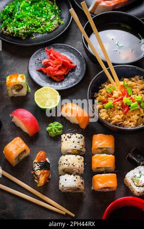 Table servie avec des sushis et de la cuisine japonaise traditionnelle sur fond sombre. Petits pains à sushi, wakame hiyashi, soupe miso, ramen, riz frit aux légumes Banque D'Images