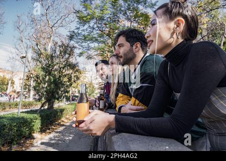 Deux hommes et deux femmes se détendent ensemble, s'appuyant sur une balustrade en pierre, tenant des boissons en bouteille (bière et soda). Vue de profil, groupe divers avec a caucasien Banque D'Images