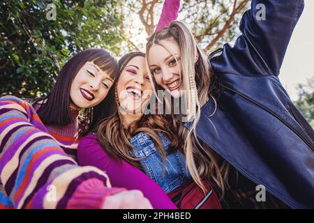 Trois jeunes femmes du Caucase rient, plaisantent, chantent et posent pour la caméra dans un parc public. Maquillage audacieux et vêtements décontractés de style 90s. Blond a Banque D'Images