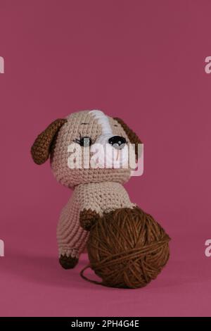 Poupée Amigurumi chiot sur fond rose jouant avec la balle de fil. Un jouet de bricolage doux en coton naturel et laine. Petit chien brun crocheté, handm Banque D'Images