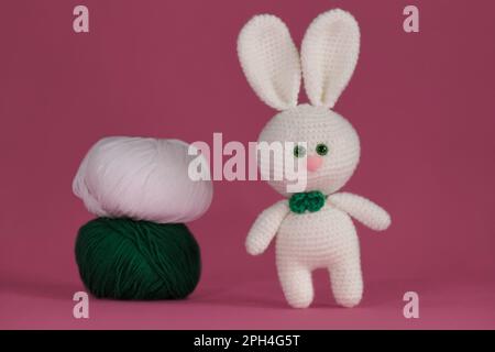 Symbole du concept de Pâques catholique. Lapin blanc de Pâques crocheté, art fait à la main Poupée de lapin Amigurumi sur fond rose avec deux boules de vert et wh Banque D'Images